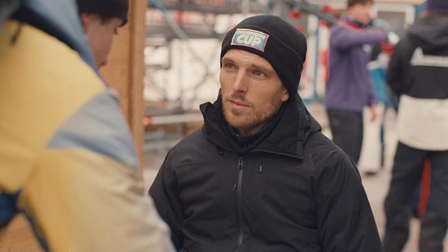 In der Mitte des Bildes ist ein junger Mann mit einer schwarzen Winterjacke und schwarzen Mütze mit der Aufschrift "Champions Cup" zu sehen, der eine vor ihm stehende Person ansieht. Im Hintergrund sind weitere Menschen zu sehen. Der junge Mann trägt einen hellen Dreitagesbart, hat blaue Augen und lächelt leicht. 
