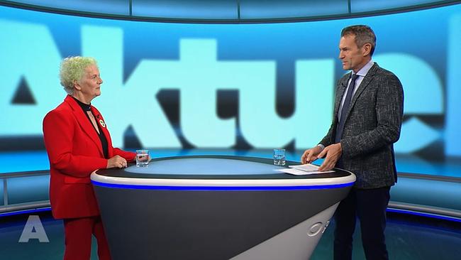 In einem Fernsehstudio steht ein Moderator rechts im Bild an einem Tisch angelehnt. Links, auf der anderen Seite des Tisches steht eine Frau, die als Interviewpartnerin eingeladen ist. Sie ist sehr auffällig gekleidet und trägt einen roten Hosenanzug. Sie hat blonde kurze Locken. Vor dem Moderator und dem Gast steht jeweils ein halb angefülltes Wasserglas. 