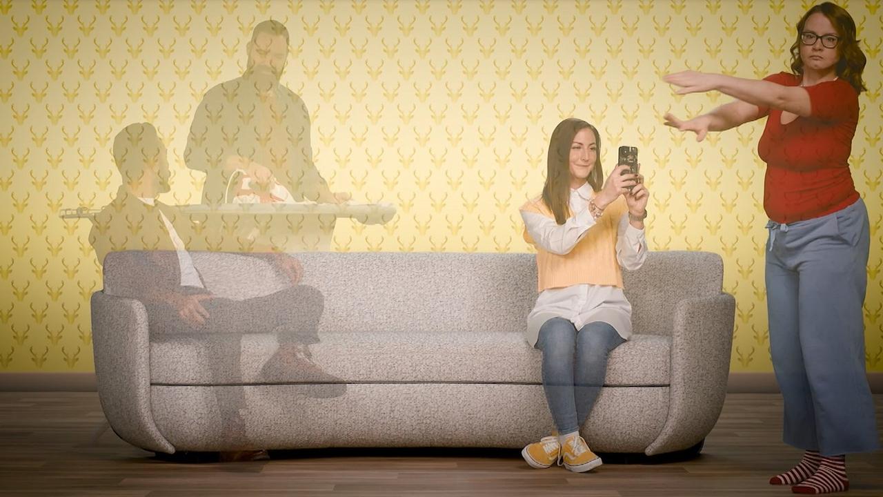 © Auf dem Bild sind vier Personen zu sehen, zwei Sitzen auf einer grauen Couch, die präsent in der Mitte des Bildes positioniert ist. Ein Mann steht hinter der Couch bei einem Bügelbrett und bügelt. Eine weitere Frau tanzt offensichtlich vor der Couch. Sie wird von einer Person, die auf der Couch sitzt, fotografiert. Es ist eine ausgelassene, fröhliche Stimmung und stellt eine Familiensituation dar.