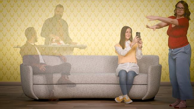 Auf dem Bild sind vier Personen zu sehen, zwei Sitzen auf einer grauen Couch, die präsent in der Mitte des Bildes positioniert ist. Ein Mann steht hinter der Couch bei einem Bügelbrett und bügelt. Eine weitere Frau tanzt offensichtlich vor der Couch. Sie wird von einer Person, die auf der Couch sitzt, fotografiert. Es ist eine ausgelassene, fröhliche Stimmung und stellt eine Familiensituation dar. 