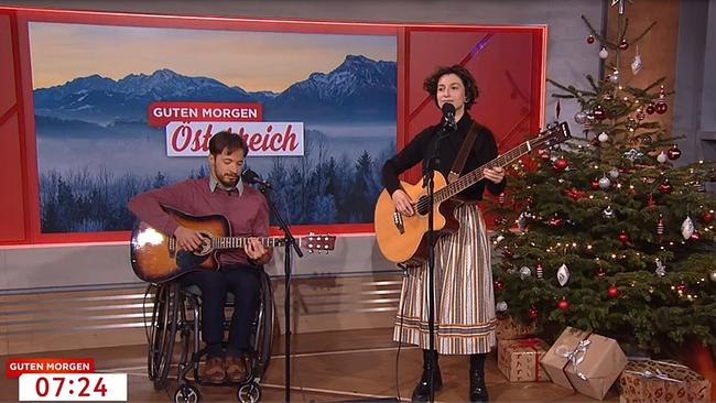 Ein junger Mann im Rollstuhl sitzt im Rollstuhl. Er spielt Gitarre und singt. Vor ihm steht ein Mikrofon. Rechts neben ihm steht eine junge Frau, ebenfalls Sängerin, die ebenso die Gitarre spielt. Hinter ihnen sieht man eine Berglandschaft, in die der Name "Guten Morgen Österreich" eingeblendet ist. In der unteren linken Ecke ist die Uhrzeit "7.24" Uhr eingeblendet. 