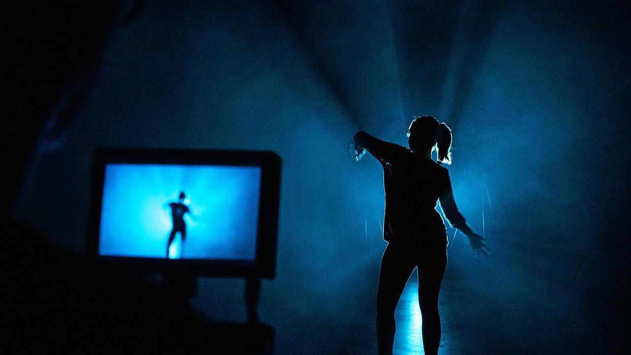 Silhouette einer Frau vor einem Bildschirm, auf dem sie zu sehen ist. Sie macht eine Tanzbewegung mit ihren Armen und trägt ihre Haare in einem Pferdeschwanz. Das Bild ist in dunkles, blaues Licht getaucht.